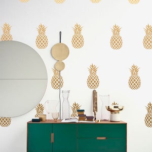May include: A green dresser with gold pineapple decals on a white wall. The dresser has two drawers and wooden legs. There are gold decorative items on top of the dresser.