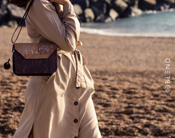 May include: A woman wearing a beige dress with a brown and white cork and purple leather crossbody bag. The bag has a strap that goes over the shoulder and a flap closure.