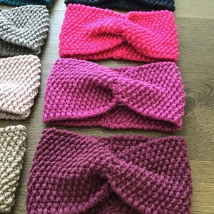 Op de afbeelding: Een verzameling gebreide hoofdbanden in verschillende kleuren, waaronder teal, zwart, grijs, roze en paars. De hoofdbanden zijn gemaakt met een dik garen en hebben een gedraaid knoopontwerp.