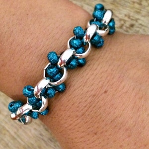 May include: A silver chain bracelet with teal blue glitter beads. The bracelet is on a person's wrist.