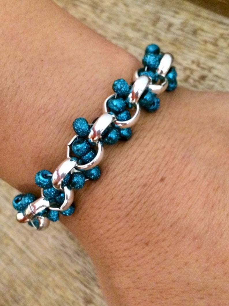 May include: A silver chain bracelet with teal blue glitter beads. The bracelet is on a person's wrist.
