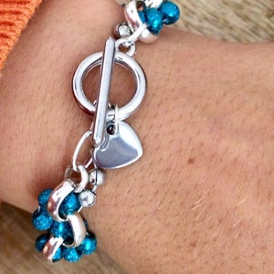 May include: A silver bracelet with a heart charm and a toggle clasp. The bracelet is adorned with turquoise blue beads.