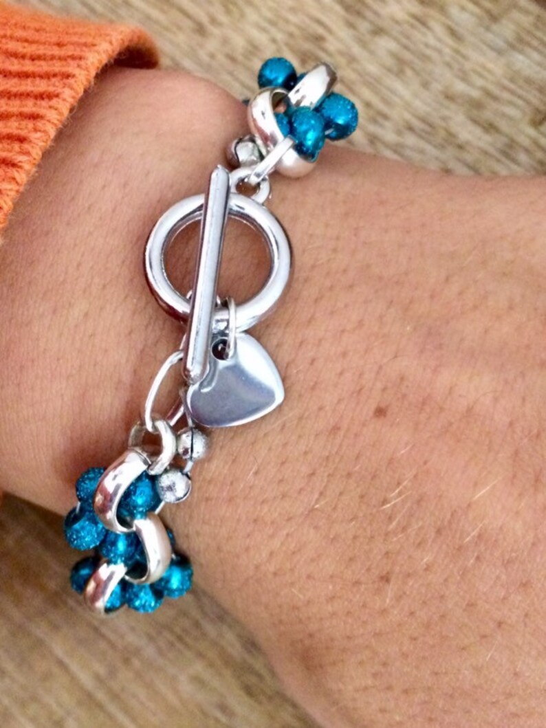 May include: A silver bracelet with a heart charm and a toggle clasp. The bracelet is adorned with turquoise blue beads.