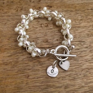 May include: A silver bracelet with a toggle clasp and two charms. One charm is a heart and the other is a round charm with the letter "O" on it. The bracelet is made of small white beads.