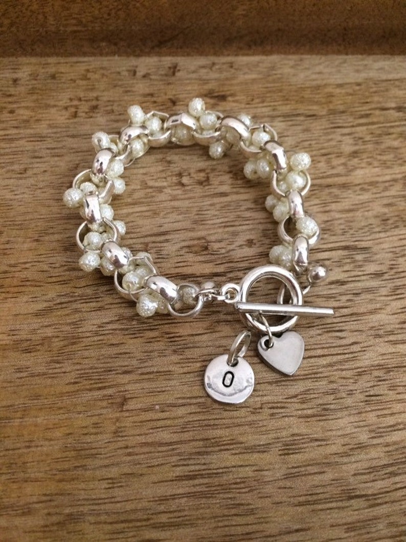 May include: A silver bracelet with a toggle clasp and two charms. One charm is a heart and the other is a round charm with the letter "O" on it. The bracelet is made of small white beads.