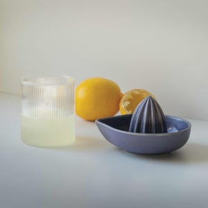 May include: A glass of freshly squeezed lemon juice in a clear glass with a ribbed design. Next to it are two lemons, one whole and one cut in half. A purple ceramic lemon squeezer sits on a white surface.