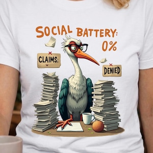May include: White t-shirt featuring a cartoon stork wearing glasses, with the text "SOCIAL BATTERY: 0%." The stork is surrounded by stacks of paper, with signs reading "CLAIMS:" and "DENIED."