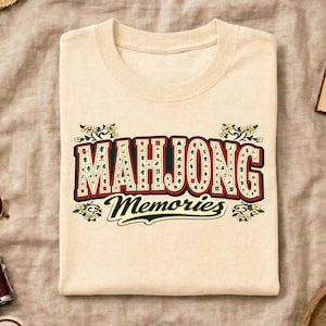 May include: A cream-colored t-shirt featuring the words "MAHJONG Memories" in a retro font. The text is in red, green, and white, with floral accents. The shirt is made of a soft material.