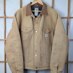 Peut inclure: Veste de travail beige avec un col en velours c&ocirc;tel&eacute; marron, une fermeture boutonn&eacute;e et deux poches poitrine. La veste a des boutons dor&eacute;s et un &eacute;cusson logo Carhartt. La veste est suspendue &agrave; un cintre en bois.