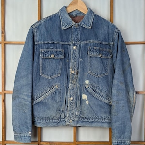 May include: A vintage blue denim jacket with a classic collar, button closure, and multiple pockets. The jacket shows signs of wear, including faded areas and paint splatters, adding to its unique, worn aesthetic. It hangs on a wooden hanger.