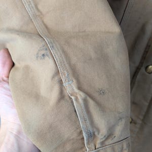 Peut inclure: Gros plan sur une manche de veste de travail beige. La veste pr&eacute;sente des signes d'usure, avec des taches et des bords effiloch&eacute;s. Des boutons de couleur laiton sont visibles. Les coutures sont pro&eacute;minentes et le tissu semble &ecirc;tre un m&eacute;lange de coton durable.