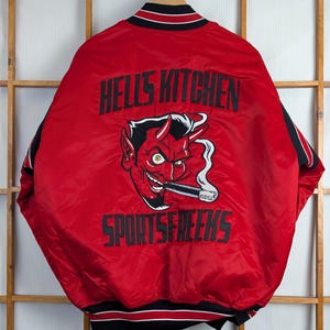 May include: Red satin bomber jacket with black and white trim. The back features a devil graphic with the text "HELL'S KITCHEN SPORTSFREEKS" in black. The jacket is hanging on a wooden background.