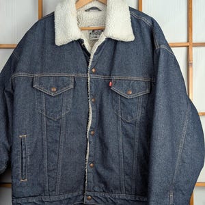 May include: A dark blue denim jacket with a white, fluffy shearling collar and lining. The jacket has two chest pockets, two side pockets, and a red Levi's tag. It is hanging on a wooden hanger against a wooden background.