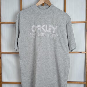 May include: A gray short-sleeved t-shirt with the white text "OAKLEY High Definition Optics" across the chest. The shirt is hanging on a wooden hanger against a wooden window frame.
