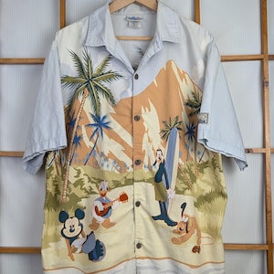May include: Button-down short-sleeve shirt with a tropical beach scene featuring Mickey Mouse, Donald Duck, Goofy, and Pluto. The shirt has a light blue collar and sleeves, with a beige and yellow body.