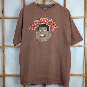 以下が含まれることがあります： 茶色のTシャツ。漫画の顔のグラフィックと「HEY HEY HEY」の文字が赤と白で描かれています。木製のハンガーに掛けられ、窓ガラスを背景にしています。