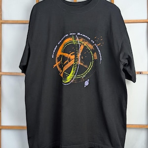 May include: Black t-shirt with a graphic design featuring a clock-like image with the text "EVERY CLOCK ON EARTH IS TICKING..." in orange, green, and white. The shirt is hanging on a wooden hanger.