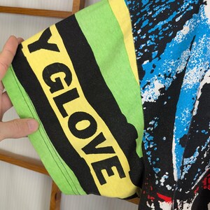 Pu&ograve; includere: Primo piano di una manica di maglietta grafica colorata. La manica presenta il testo "Y GLOVE" in grandi lettere gialle su uno sfondo a righe nere e verdi. Il design include schizzi di vernice blu e bianca.