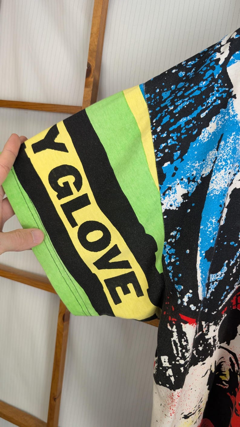 Pu&ograve; includere: Primo piano di una manica di maglietta grafica colorata. La manica presenta il testo "Y GLOVE" in grandi lettere gialle su uno sfondo a righe nere e verdi. Il design include schizzi di vernice blu e bianca.