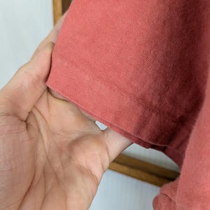Puede incluir: Primer plano de una manga de camiseta de color coral. La tela parece ser un tejido de punto suave. La manga est&aacute; sujeta por una mano, mostrando el dobladillo y la textura del material. El fondo es de color claro.