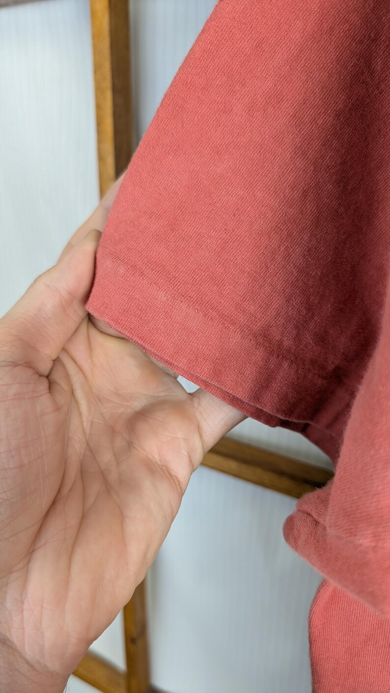 Puede incluir: Primer plano de una manga de camiseta de color coral. La tela parece ser un tejido de punto suave. La manga est&aacute; sujeta por una mano, mostrando el dobladillo y la textura del material. El fondo es de color claro.