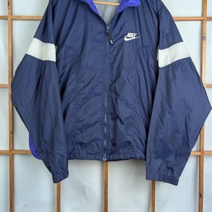 Op de afbeelding: Marineblauwe Nike windjack met witte accenten op de mouwen en een paarse kraag. De jas heeft een volledige ritssluiting en het Nike-logo op de borst. Het hangt aan een houten hanger.