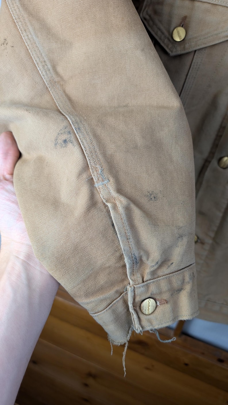 Peut inclure: Gros plan sur une manche de veste de travail beige. La veste pr&eacute;sente des signes d'usure, avec des taches et des bords effiloch&eacute;s. Des boutons de couleur laiton sont visibles. Les coutures sont pro&eacute;minentes et le tissu semble &ecirc;tre un m&eacute;lange de coton durable.