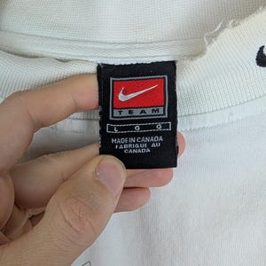 Peut inclure: Sweat-shirt cr&egrave;me Nike Team avec une &eacute;tiquette noire et rouge. L'&eacute;tiquette indique "MADE IN CANADA". Le sweat-shirt pr&eacute;sente un motif graphique avec des accents orange et rouge.