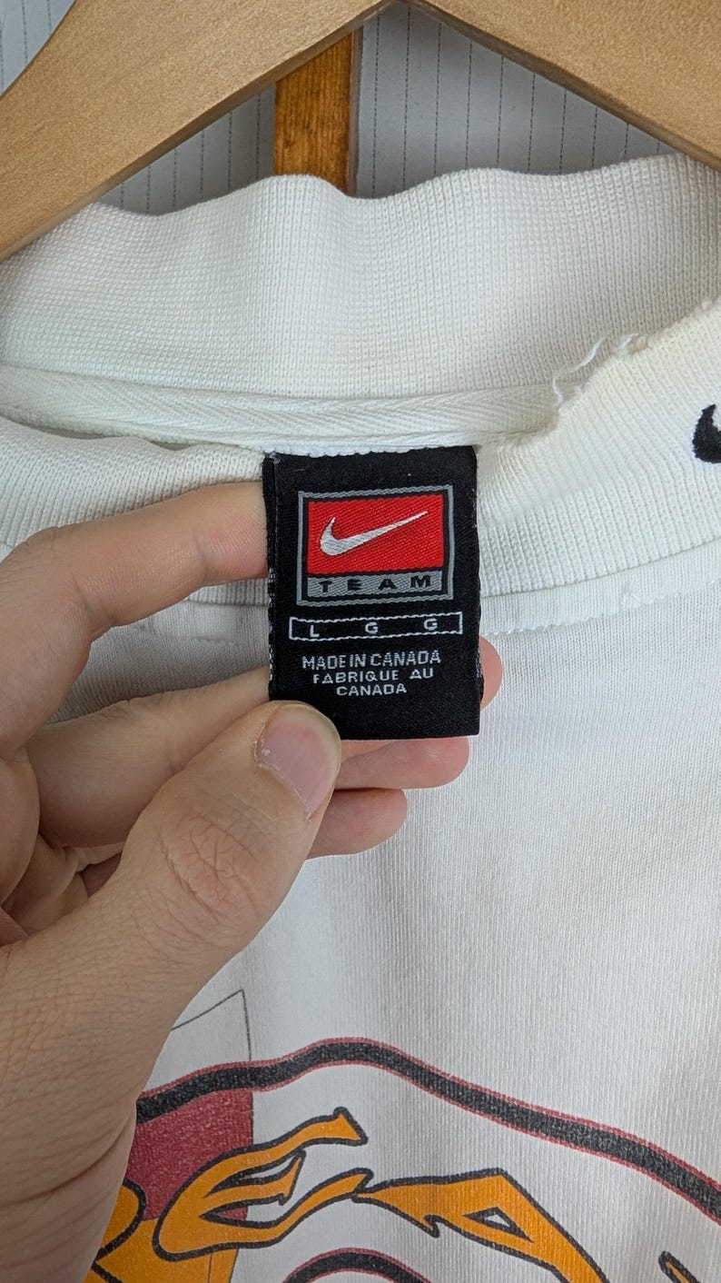 Peut inclure: Sweat-shirt cr&egrave;me Nike Team avec une &eacute;tiquette noire et rouge. L'&eacute;tiquette indique "MADE IN CANADA". Le sweat-shirt pr&eacute;sente un motif graphique avec des accents orange et rouge.