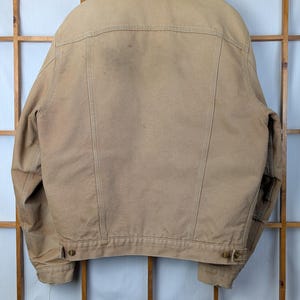 Peut inclure: Veste de travail beige avec un col en velours c&ocirc;tel&eacute; marron suspendue &agrave; un cintre en bois. Le dos de la veste est visible, montrant les coutures et les fermetures &agrave; boutons. La veste est faite d'un mat&eacute;riau durable, adapt&eacute; au travail en ext&eacute;rieur.