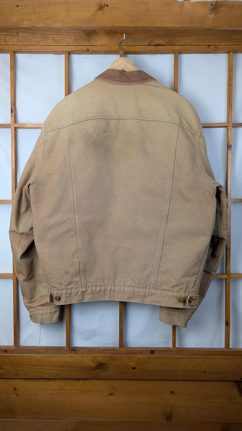 Peut inclure: Veste de travail beige avec un col en velours c&ocirc;tel&eacute; marron suspendue &agrave; un cintre en bois. Le dos de la veste est visible, montrant les coutures et les fermetures &agrave; boutons. La veste est faite d'un mat&eacute;riau durable, adapt&eacute; au travail en ext&eacute;rieur.