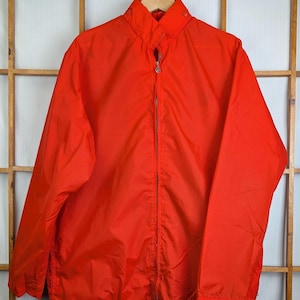 May include: Bright red jacket with a full zipper closure and a stand-up collar. The jacket is hanging on a wooden hanger. The jacket has long sleeves and a drawstring at the bottom hem. The jacket is made of a lightweight material.