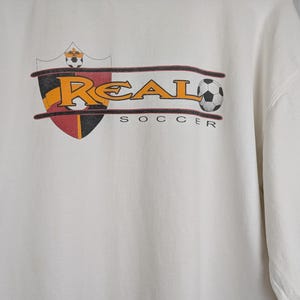 Peut inclure: Sweat-shirt blanc &agrave; manches longues avec un col montant et un logo Nike noir. Le devant pr&eacute;sente un graphisme sur le th&egrave;me du football avec le mot "REAL" en lettres dor&eacute;es, un ballon de football et le mot "SOCCER".
