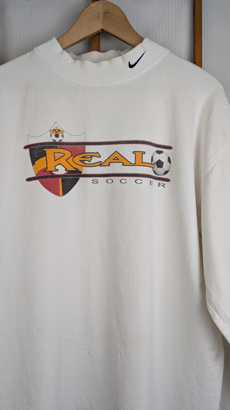 Peut inclure: Sweat-shirt blanc &agrave; manches longues avec un col montant et un logo Nike noir. Le devant pr&eacute;sente un graphisme sur le th&egrave;me du football avec le mot "REAL" en lettres dor&eacute;es, un ballon de football et le mot "SOCCER".