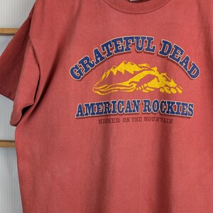 Puede incluir: Una camiseta de color rojo anaranjado con las palabras "Grateful Dead" en azul arqueadas en la parte superior, y "American Rockies" en azul debajo de un gr&aacute;fico de monta&ntilde;a amarilla. El texto "Higher on the Mountain" est&aacute; debajo.