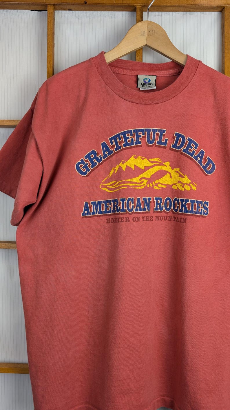 Puede incluir: Una camiseta de color rojo anaranjado con las palabras "Grateful Dead" en azul arqueadas en la parte superior, y "American Rockies" en azul debajo de un gr&aacute;fico de monta&ntilde;a amarilla. El texto "Higher on the Mountain" est&aacute; debajo.