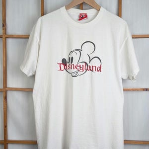 Puede incluir: Camiseta blanca con un dise&ntilde;o de Disneyland en negro y rojo. El dise&ntilde;o incluye un contorno de Mickey Mouse y la palabra "Disneyland" en una fuente estilizada. La camiseta est&aacute; colgada en una percha de madera.