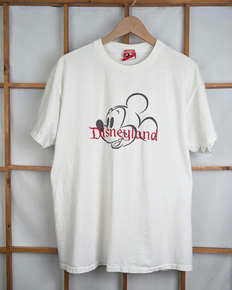 Puede incluir: Camiseta blanca con un dise&ntilde;o de Disneyland en negro y rojo. El dise&ntilde;o incluye un contorno de Mickey Mouse y la palabra "Disneyland" en una fuente estilizada. La camiseta est&aacute; colgada en una percha de madera.
