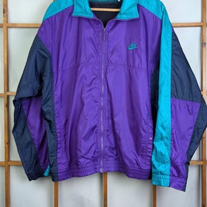 Könnte beinhalten: Eine Vintage Nike Windjacke in Lila, Türkis und Marineblau. Die Jacke hat einen durchgehenden Reißverschluss, ein kleines Nike-Logo auf der Brust und hängt an einem Holzbügel. Die Jacke ist aus einem leichten Material, möglicherweise Nylon.