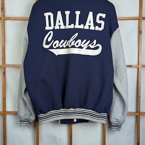 Peut inclure: Veste de style universitaire bleu marine et grise avec l'inscription "DALLAS Cowboys" en lettres blanches au dos. La veste a des manches grises, un col rayé et un ourlet rayé. La veste est suspendue à un cintre en bois.