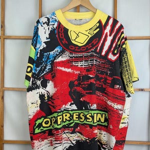 Pu&ograve; includere: T-shirt grafico vintage con colletto e maniche gialle. La maglietta presenta un design audace e colorato con le parole "OPPRESSIN" e "BODY GLOVE" in un font in stile graffiti. Il design incorpora elementi rossi, neri, blu e bianchi.