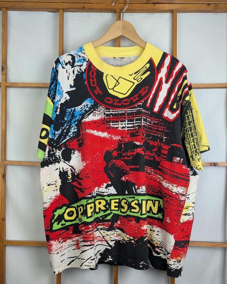 Pu&ograve; includere: T-shirt grafico vintage con colletto e maniche gialle. La maglietta presenta un design audace e colorato con le parole "OPPRESSIN" e "BODY GLOVE" in un font in stile graffiti. Il design incorpora elementi rossi, neri, blu e bianchi.
