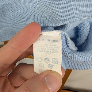May include: Close-up of a pale blue garment with a label stating "MADE IN USA". The label also includes washing instructions. The fabric has a ribbed texture.