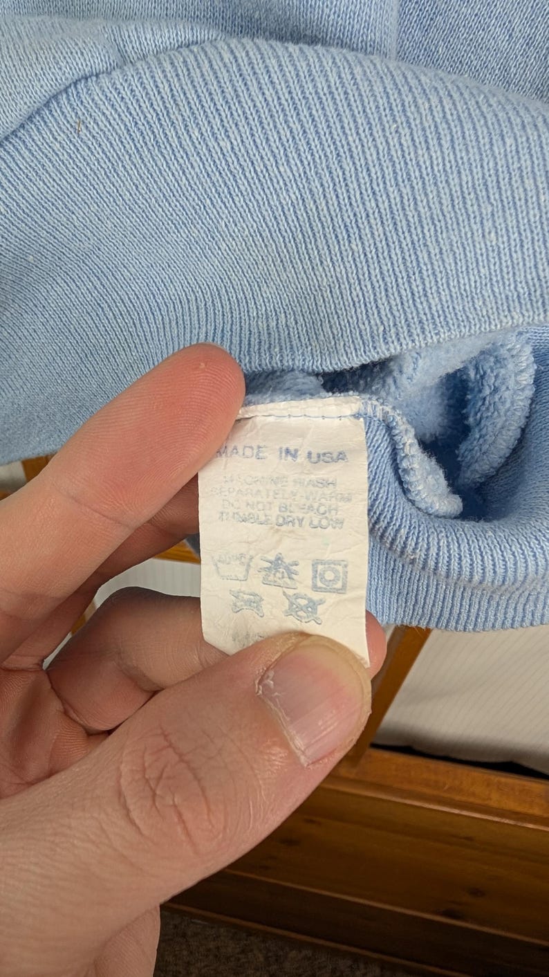 May include: Close-up of a pale blue garment with a label stating "MADE IN USA". The label also includes washing instructions. The fabric has a ribbed texture.