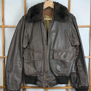 May include: A brown leather bomber jacket with a dark brown fur collar. The jacket features two front pockets with button closures, a zip closure, and ribbed cuffs and hem. It is suspended on a wooden hanger.
