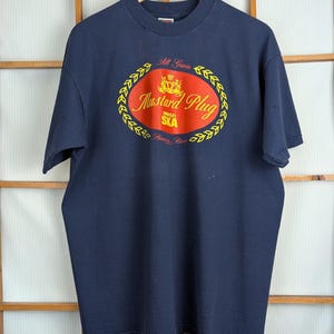May include: Navy blue t-shirt with a yellow and red oval design. The design features the words "Mustard Plug" and "SKA" in a stylized font, with the words "All Grain" above and "Brewery Fresh" below. The shirt is hanging on a wooden hanger.