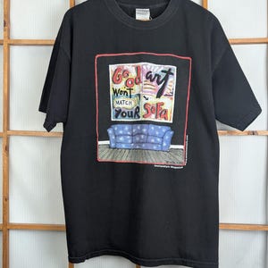 May include: Black t-shirt with a colorful graphic. The graphic features the text "Good art won't match your sofa" above a blue patterned sofa. The shirt is hanging on a wooden hanger.
