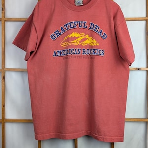 Puede incluir: Una camiseta roja con el texto "Grateful Dead American Rockies" en azul y amarillo. La camiseta est&aacute; colgada en una percha de madera contra un marco de ventana de madera.