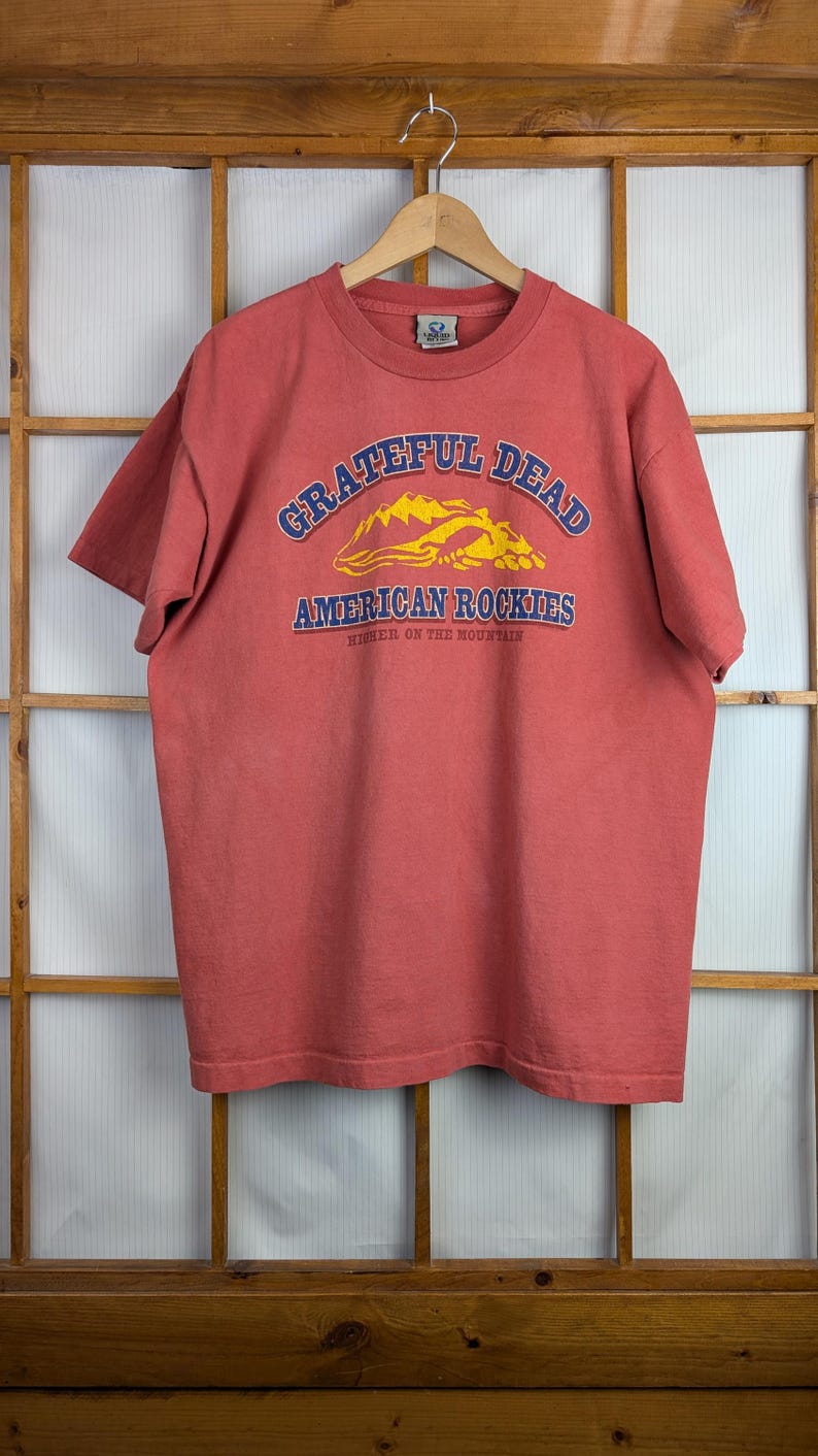 Puede incluir: Una camiseta roja con el texto "Grateful Dead American Rockies" en azul y amarillo. La camiseta est&aacute; colgada en una percha de madera contra un marco de ventana de madera.