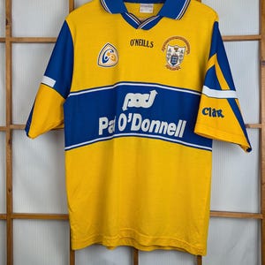 Może przedstawiać: Żółto-niebieska koszulka O'Neills z tekstem Pa O'Donnell. Koszulka ma niebieski kołnierz i rękawy z białymi akcentami. Koszulka wisi na drewnianym wieszaku na drewnianym tle.